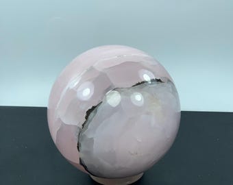 Könnte beinhalten: Eine polierte rosa Onyx-Kugel mit dunkelgrauer Maserung. Die Kugel hat eine glatte, reflektierende Oberfläche und steht auf einem kleinen, kreisförmigen Sockel. Die Farbe des Steins variiert von hell- bis mittelrosa.