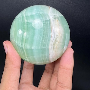May include: A polished, spherical green onyx stone featuring natural banded patterns in shades of light mint, sage, and creamy white. The surface is smooth and reflective, showcasing translucent layers and organic mineral striations throughout the orb.