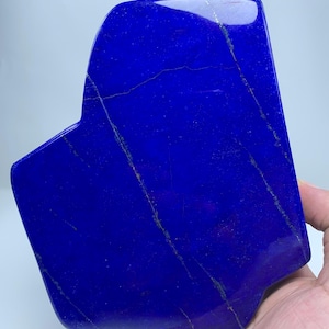 Op de afbeelding: Een gepolijste, diepblauwe lapis lazuli plaat met een glad, reflecterend oppervlak. De steen heeft opvallende, dunne, diagonale goudkleurige pyrietaders die over het oppervlak lopen en een natuurlijk patroon in het mineraal vormen.