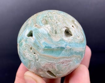Puede incluir: Una gema esférica pulida, con un patrón arremolinado de tonos azules, verdes y beige. La piedra tiene varios agujeros pequeños y naturales y se sostiene en una mano sobre un fondo oscuro. Es un objeto decorativo natural.