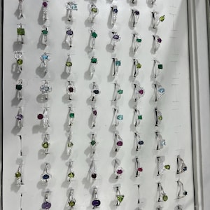 Könnte beinhalten: Eine Präsentation von Silberringen mit verschiedenen Edelsteinen, darunter Smaragde, Amethyste und Rubine. Die Ringe sind in einem weißen Display angeordnet und zeigen eine Vielzahl von Designs und Steinschnitten. Die Edelsteine haben verschiedene Formen.