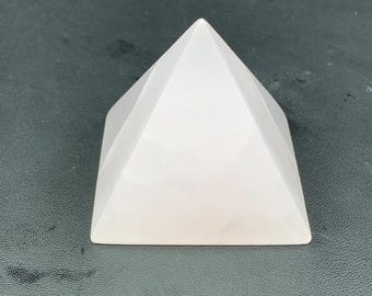 Op de afbeelding: Een lichtroze, facetgeslepen piramide gemaakt van een doorschijnend materiaal. De piramide heeft een platte basis en een puntige top. Het object staat op een donkere ondergrond.