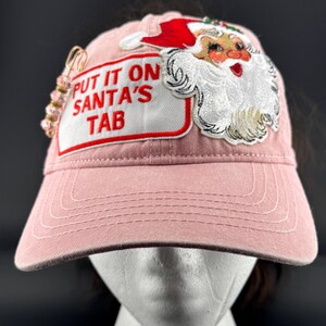 May include: A dusty pink baseball cap featuring an embroidered Santa Claus patch and a white rectangular patch with the red text "PUT IT ON SANTA'S TAB". A gold-toned safety pin adorned with small pink beads is attached to the side of the cap.