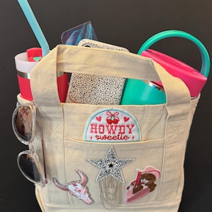 May include: A beige canvas tote bag with a "Howdy Sweetie" patch, star and cowgirl patches, and sunglasses. The bag contains a tumbler, a pink container, a phone case, and a blue straw. The bag has a handle.