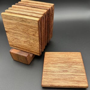 Può includere: Set di sottobicchieri in legno con una pila di sottobicchieri quadrati e una piccola base rettangolare. I sottobicchieri sono in legno marrone chiaro con venature visibili. Lo sfondo è nero.