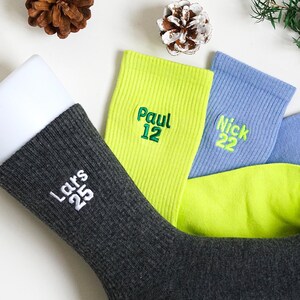 May include: A selection of personalised socks in different colours. One dark grey sock with "Lars 25" embroidered in white is on a white form. Other socks are lime green, light blue, and blue, each with a name and number in green.