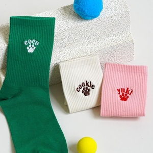 Könnte beinhalten: Grüne, cremefarbene und rosa Socken mit Pfotenabdruck-Designs. Die grüne Socke hat die Stickerei "coco", die cremefarbene Socke "cookie" und die rosa Socke "yuki". Ein blauer Pompon und ein gelber Ball sind ebenfalls im Bild.