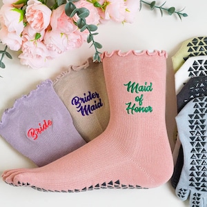 May include: A collection of wedding-themed socks in various colors, including pink, lavender, and tan. The socks feature text like "Bride," "Bridesmaid," and "Maid of Honor." Non-slip socks with a toe design are also visible.