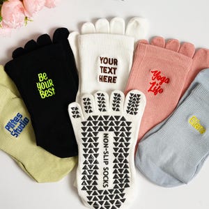 Op de afbeelding: Diverse teensokken in verschillende kleuren, waaronder zwart, wit, roze en lichtblauw. Sommige sokken hebben teksten als "Be your Best", "Yoga Life" en "Elite". Eén witte sok heeft een antislipzoolontwerp.