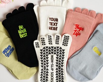 Op de afbeelding: Diverse teensokken in verschillende kleuren, waaronder zwart, wit, roze en lichtblauw. Sommige sokken hebben teksten als "Be your Best", "Yoga Life" en "Elite". Eén witte sok heeft een antislipzoolontwerp.