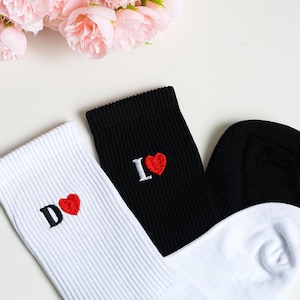 Könnte beinhalten: Drei Paar Socken, eine schwarze und zwei weiße, mit gesticktem Text und einem roten Herz. Die schwarze Socke sagt "I ❤️" und die weißen Socken sagen "D ❤️". Rosa Blumen sind im Hintergrund.