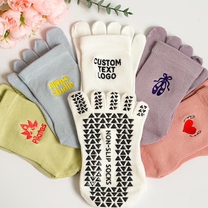 Könnte beinhalten: Verschiedene Zehensocken in Pastellfarben, darunter Weiß, Lavendel und Hellgrün. Einige Socken haben gestickte Designs wie ein Herz, einen Ballettschuh und Text. Ein Paar hat "CUSTOM TEXT LOGO" aufgedruckt, ein anderes "NON-SLIP SOCKS" auf der Sohle.
