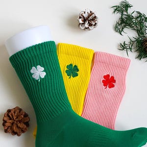 May include: Three pairs of socks in green, yellow, and pink, each featuring a four-leaf clover motif. The green sock has a white clover, the yellow a green clover, and the pink a red clover. The socks are displayed on a white surface.