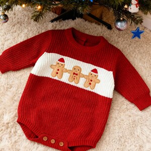 Op de afbeelding: Rode gebreide babyromper met lange mouwen. De romper heeft een witte horizontale streep over de borst met drie gingerbread man applicaties met kerstmutsen. De romper heeft drie houten knopen aan de onderkant.