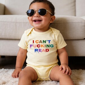 May include: A baby wearing a light yellow onesie and round sunglasses, smiling. The onesie features the text "I CAN'T FUCKING READ" in rainbow colours with a dripping paint effect. The baby is seated in front of a beige sofa.