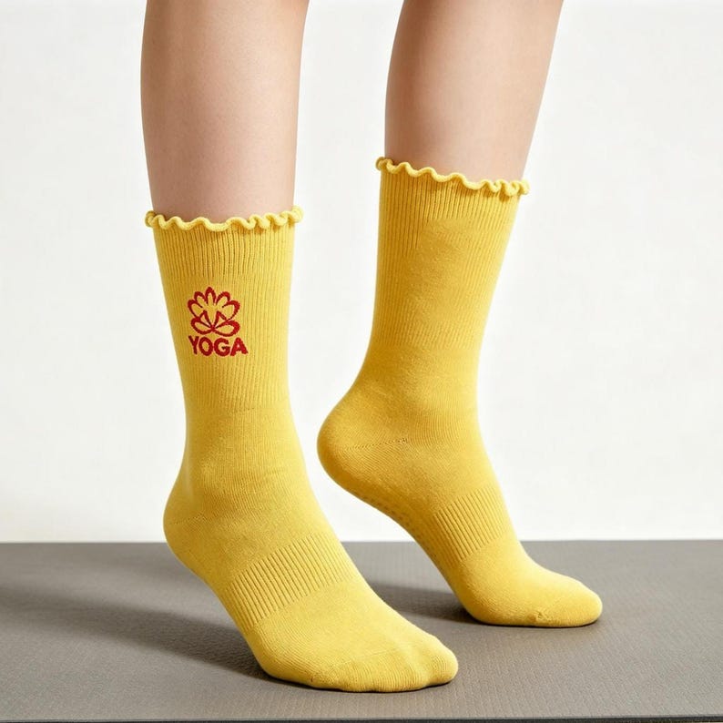 May include: A pair of mustard yellow crew socks featuring a ruffled lettuce-edge cuff. The right sock displays a red embroidered lotus flower icon above the word YOGA in a clean, sans-serif font. The socks have a ribbed texture along the leg.