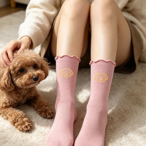 May include: A pair of dusty pink ribbed socks featuring a ruffled lettuce-edge cuff and a small, yellow embroidered outline of a curly-haired dog face on the front of each sock, positioned mid-calf on the lower legs.