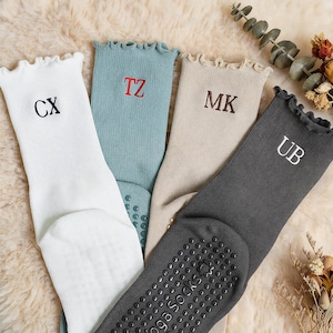 Op de afbeelding: Vier paar geribbelde yogasokken met ruches aan de boorden in wit, blauwgroen, beige en donkergrijs. Elke sok heeft geborduurde initialen: CX in zwart, TZ in rood, MK in bruin en UB in wit. De zolen zijn voorzien van zwarte siliconen antislipstippen.