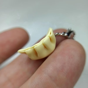 Può includere: Un piccolo ciondolo a forma di raviolo giallo pallido con pieghe dettagliate, attaccato a una catena color argento. Il raviolo è tenuto tra le dita, evidenziando le sue piccole dimensioni e l'aspetto realistico. Un accessorio a tema cibo.