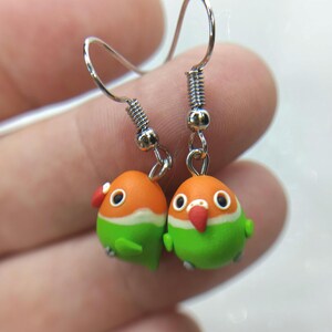Puede incluir: Un par de pendientes hechos a mano con forma de pájaro. Cada pendiente presenta una pequeña cabeza redonda naranja con una franja blanca, un cuerpo verde y un pico rojo. Los pendientes están suspendidos de ganchos plateados.