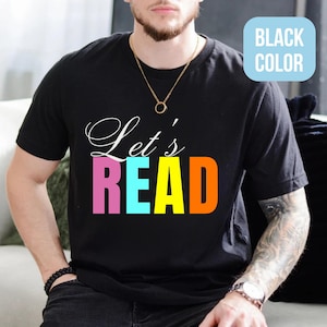 May include: Black t-shirt featuring the words "Let's READ" in white, pink, blue, and orange. The shirt has a crew neck and short sleeves. The words "BLACK COLOR" are in a light blue box in the upper right corner.