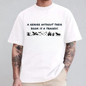 Puede incluir: Una camiseta blanca de manga corta con el texto negro A READER WITHOUT THEIR BOOK IS A TRAGEDY. Debajo del texto hay una fila de siluetas negras que incluyen un hada, un dragón, murciélagos, estrellas, una luna creciente, un castillo, un mago y un unicornio.