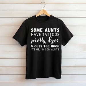 Op de afbeelding: Een zwart t-shirt met korte mouwen en witte tekst: SOME AUNTS HAVE TATTOOS pretty Eyes & CUSS TOO MUCH IT'S ME, I'M SOM AUNTS. Het t-shirt hangt aan een houten kledinghanger tegen een achtergrond van horizontale witte panelen.