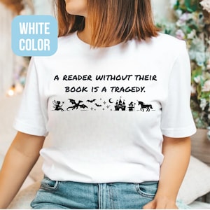 Op de afbeelding: Wit T-shirt met de tekst "A reader without their book is a tragedy." Onder de tekst staat een zwart silhouetontwerp van een fee, draak, vleermuizen, sterren, maan, kasteel, eenhoorn en een persoon.