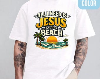 May include: White t-shirt with a colorful graphic. The design features the text "ALL I NEED IS JESUS AND THE BEACH" in yellow and black, with a beach scene including palm trees, waves, and a sunset.