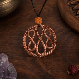 Op de afbeelding: Een ronde hanger gemaakt van gedraaid koperdraad dat een ingewikkeld oneindigheidsknoopmotief vormt. De hanger hangt aan een zwart koord en heeft een kleine, getextureerde, oranjebruine bolvormige kraal boven het metalen ontwerp.