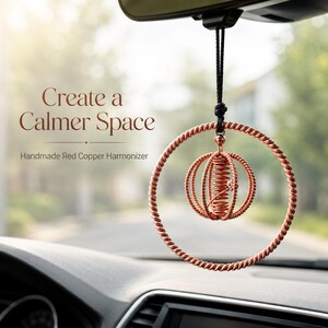 Op de afbeelding: Een roodkoperen hangend ornament met een gedraaid cirkelvormig frame en een centrale bolvormige draadkooistructuur, opgehangen aan een zwart koord. De tekst "Create a Calmer Space" en "Handmade Red Copper Harmonizer" is zichtbaar op de achtergrond.