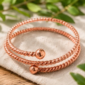 Puede incluir: Un brazalete de metal color cobre con un diseño de doble vuelta y una textura de cuerda trenzada. La pulsera termina en dos cuentas esféricas lisas y pulidas en cada extremo, creando una forma de espiral continua.