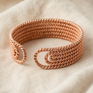 May include: A copper-colored cuff bracelet constructed from multiple parallel strands of twisted metal wire. The design features open ends that curve inward into circular loops, creating a continuous, textured, and layered appearance throughout.