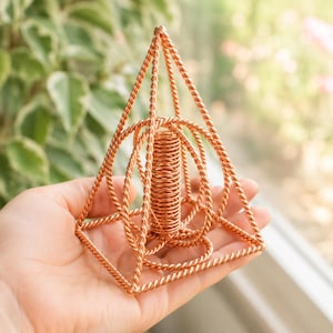 May include: A geometric sculpture constructed from twisted copper wire, featuring a pyramid frame enclosing a central cylindrical coil and circular wire elements. The structure is composed of interconnected metallic strands forming a complex, open-work design.
