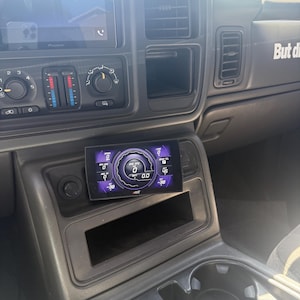 May include: A black car interior with a digital gauge display showing a speed of 0 mph and a RPM of 0. The gauge is mounted in the center console below the radio.