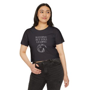 May include: Black crop top with the text "BLEEDING BUT STILL LEADING" in white. Below the text is a white crescent moon design with stars and a figure. The shirt is short-sleeved and made of a soft material.