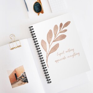May include: A spiral-bound notebook with a watercolor illustration of a branch with leaves in shades of brown. The text "Expect nothing appreciate everything" is written in a matching brown font. Gold-toned sunglasses and a binder clip are also visible.