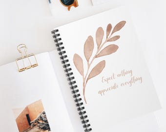 May include: A spiral-bound notebook with a watercolor illustration of a branch with leaves in shades of brown. The text "Expect nothing appreciate everything" is written in a matching brown font. Gold-toned sunglasses and a binder clip are also visible.