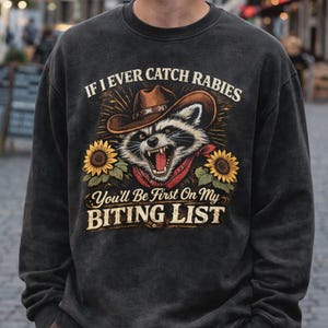 Peut inclure: Sweat-shirt gris foncé avec un raton laveur portant un chapeau de cowboy et un bandana rouge. Le motif comprend des tournesols et le texte "If I Ever Catch Rabies, You'll Be First On My Biting List."