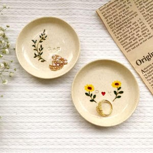 Puede incluir: Dos pequeños platos de cerámica moteada para anillos. Uno presenta un diseño de hojas verdes con las letras "G+F" y joyas doradas. El otro tiene dos girasoles amarillos, un corazón rojo, las letras "D+K" y un anillo dorado.