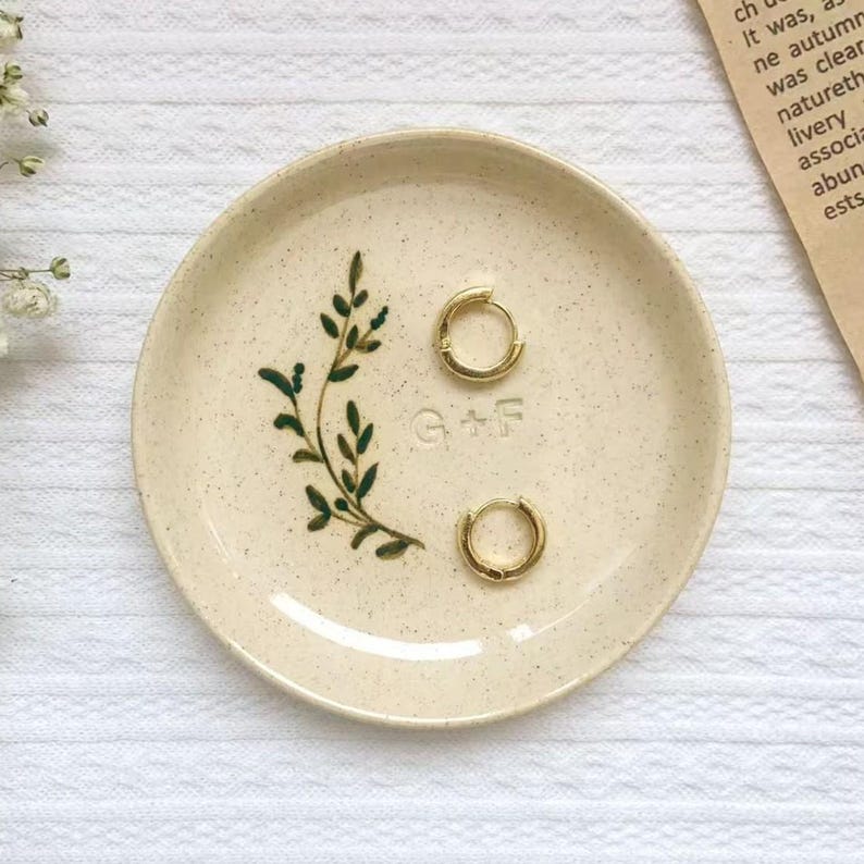 May include: A small, speckled ceramic dish with a hand-painted green leaf design and the initials "G+F". Two gold hoop earrings rest on the dish. The dish is beige and sits on a white surface.