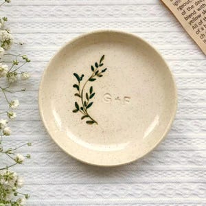 May include: A small, speckled ceramic dish with a cream-colored base. It features a dark green painted botanical design and the initials "G+F" embossed on the surface. The dish is round and has a slightly raised edge.