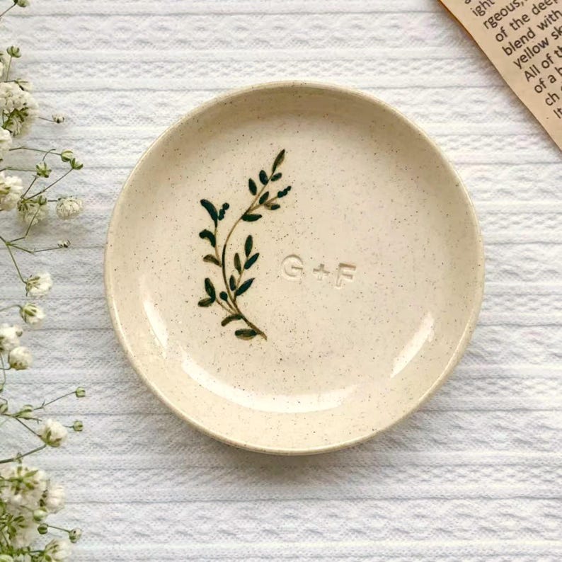 May include: A small, speckled ceramic dish with a cream-colored base. It features a dark green painted botanical design and the initials "G+F" embossed on the surface. The dish is round and has a slightly raised edge.
