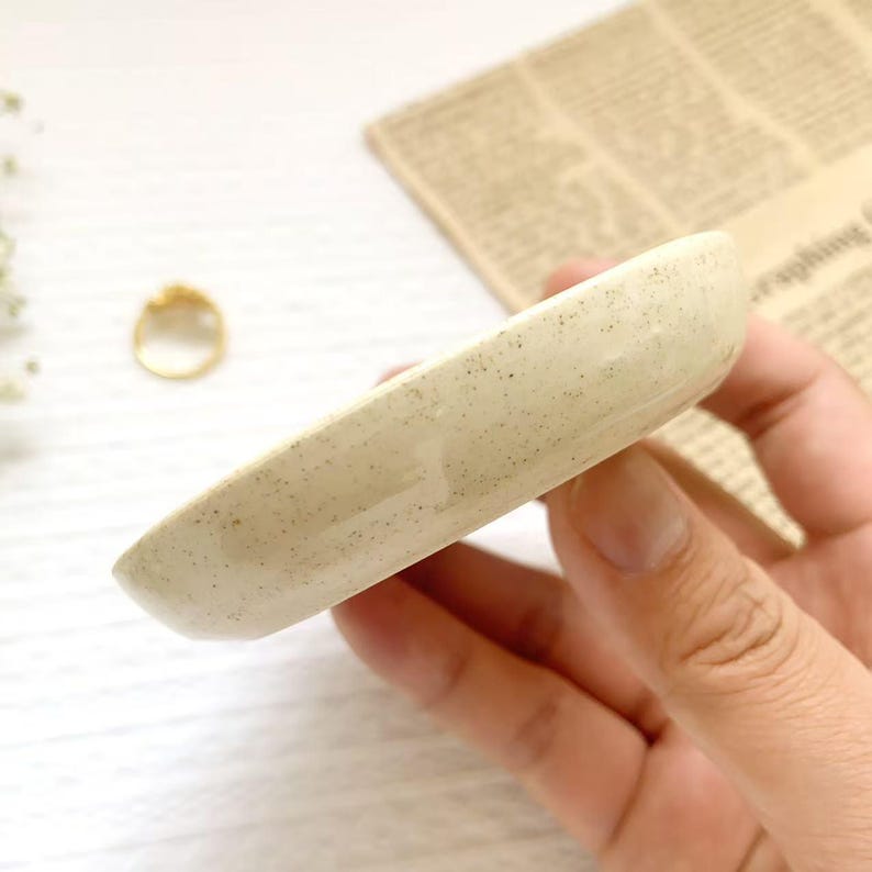 May include: A small, speckled, off-white ceramic dish, likely for jewelry or small items. The dish is oval-shaped with slightly raised edges. A gold ring is visible in the background, along with a piece of newspaper.