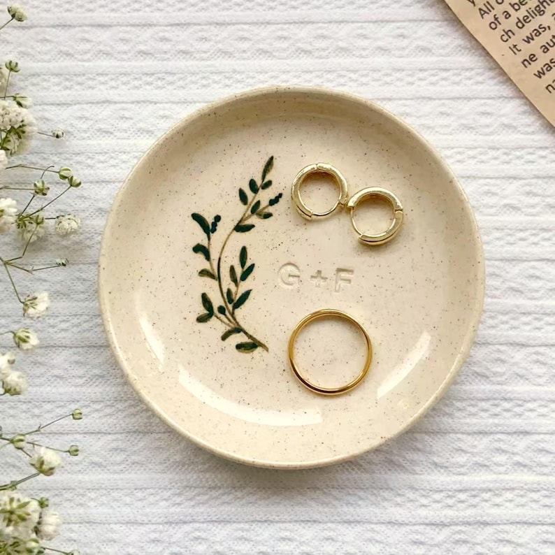May include: A small, speckled ceramic ring dish with a hand-painted green leaf design and the embossed letters "G+F". Two gold hoop earrings and a gold ring rest on the dish.