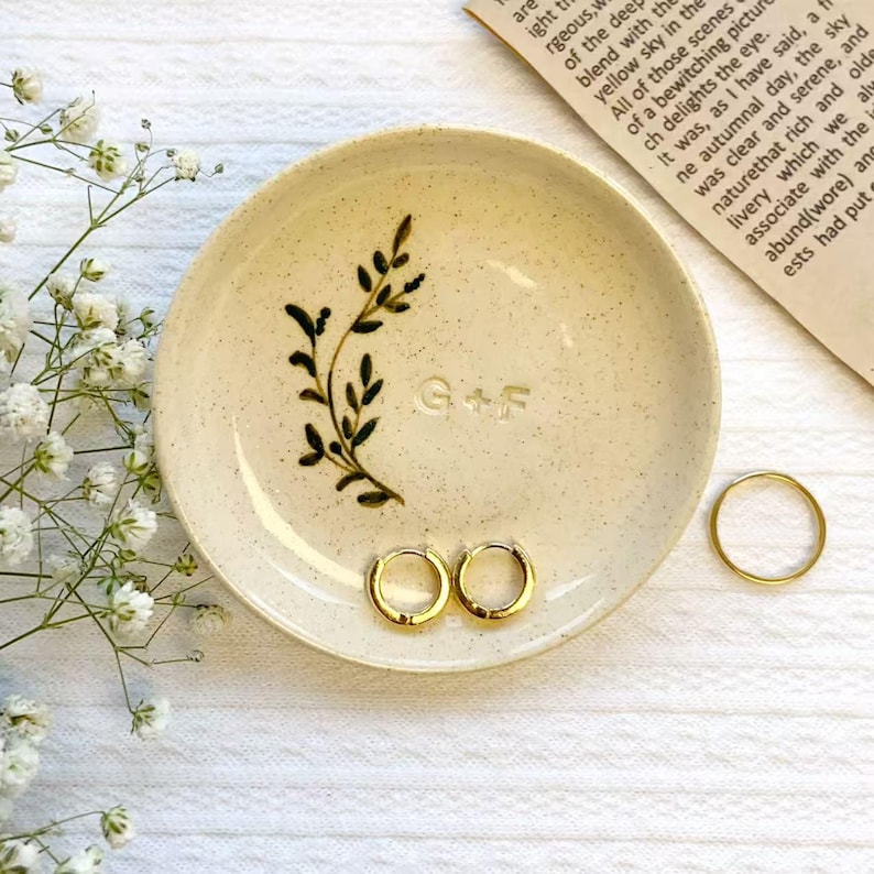 May include: A cream-colored ceramic ring dish with a hand-painted black leaf design and the initials "G+F". Two gold hoop earrings and a gold ring rest on the dish.