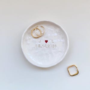 May include: A white ceramic ring dish with a textured surface. The dish features the initials "J" and "L" with a red heart symbol, the date "02.14.2026", and two gold rings. A gold square ring is also present.