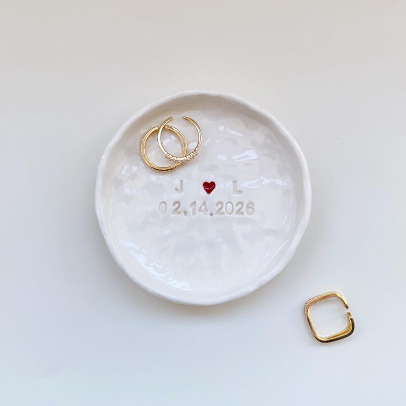 May include: A white ceramic ring dish with a textured surface. The dish features the initials "J" and "L" with a red heart symbol, the date "02.14.2026", and two gold rings. A gold square ring is also present.