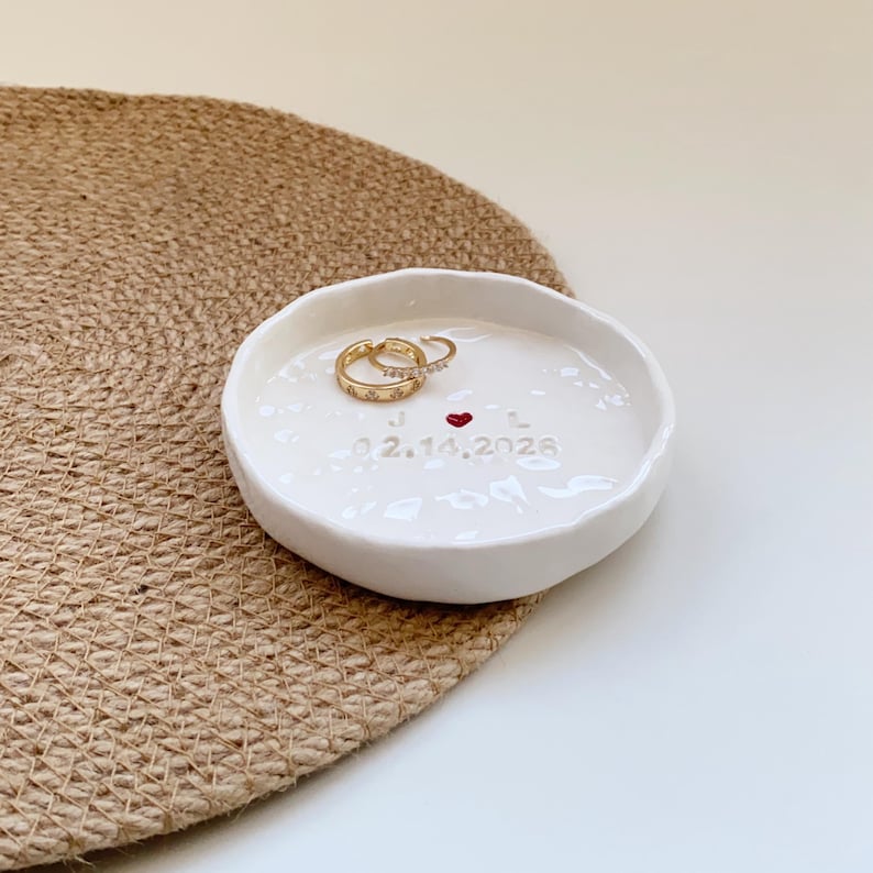 May include: A white ceramic ring dish with a slightly irregular shape. Two gold rings with clear stones rest inside. The dish has a red heart and the date 02.14.2026. The dish sits on a woven, tan placemat.