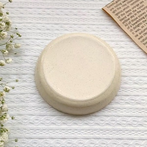 May include: A small, round, off-white ceramic dish with a speckled surface. The dish has a slightly raised edge and sits on a white, textured surface. The dish is likely used for holding small items or as a decorative piece.
